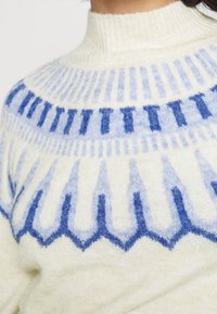 Close-up of a cream sweater with blue geometric patterns and a ribbed high neck collar on a person.