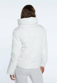 Chaqueta de plumas blanca con cuello alto, con costuras en canal y una textura suave, llevada sobre pantalones de chándal grises.