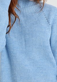 Maglione lavorato a maglia blu chiaro con colletto alto, caratterizzato da una texture a costine e un dettaglio decorativo sulla spalla.