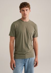 Camiseta de color verde oliva, de manga corta y fabricada en una tela suave, con un clásico cuello redondo y un corte relajado. Se lleva con unos jeans de color azul claro.