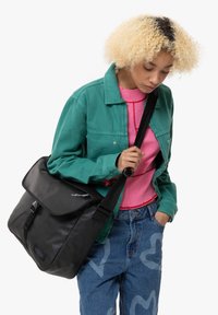 Black messenger bag with a flap, adjustable strap, and zipper detail, worn with a teal jacket and pink sweater. Denim jeans feature heart patterns.