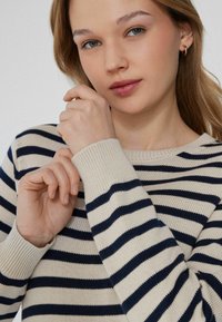 Beżowy sweter w granatowo-beżowe paski z prążkowanym dekoltem i mankietami, miękka dzianinowa struktura, dopasowany krój oraz długie rękawy.