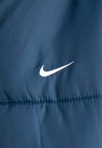 Mornarska puhovka z gladkim blagom in horizontalnim šivom; ima belo vezeno Nike logo spredaj.