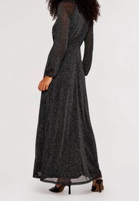 Pitkä musta iltapuku läpinäkyvinä pitkinä hihoina, laskeutuva siluetti ja hienovarainen kimaltava tekstuuri. Yhdistetty korkeakorkoisiin kenkiin.