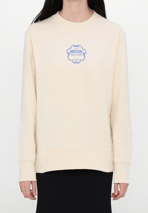 Person trägt einen beigen Pullover mit kleinem blauem Moschino Jeans Logo auf der Brust, kombiniert mit einem schwarzen Rock.