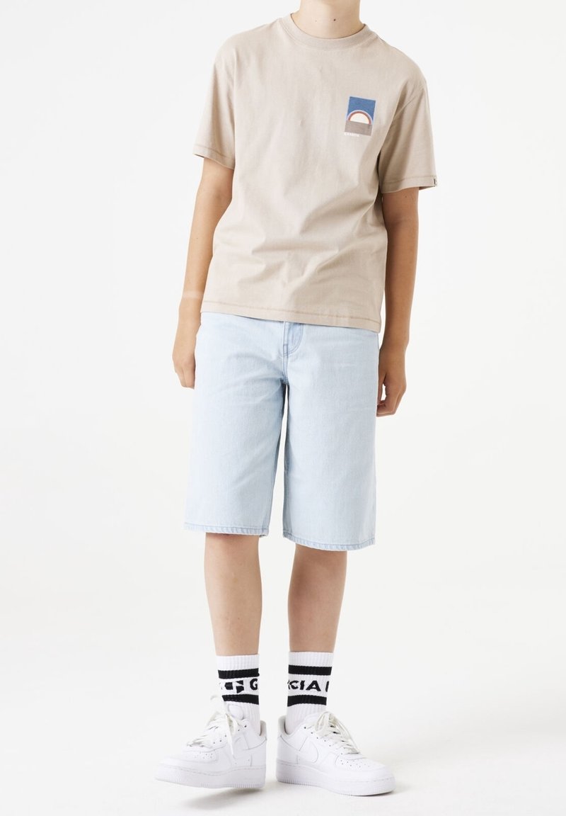 Personne portant un t-shirt beige à manches courtes avec un motif graphique, un short en jean bleu clair, des baskets blanches et des chaussettes blanches à rayures noires avec du texte.