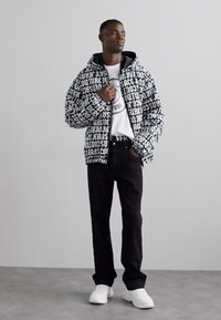 Veste zippée noir et blanc avec un grand motif textuel, portée sur un t-shirt graphique blanc. Un pantalon noir slim et des baskets blanches complètent le look.