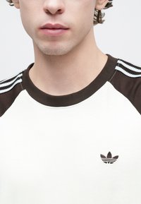 Λευκό βαμβακερό μπλουζάκι με καφέ μανίκια ράγκλαν, ριμπ λαιμόκοψη και κεντημένο λογότυπο με τρεις ρίγες στους ώμους.