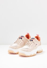 Sneaker beige con una finitura liscia, interno arancione, suola bianca e suola traslucida. Presenta lacci e un logo sulla linguetta.