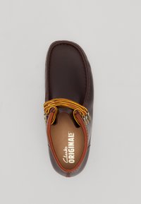 Zapato de cuero marrón con punta redonda, que presenta un acento de cordones multicolores trenzados y herrajes decorativos. El interior muestra el logotipo de Clarks Originals.