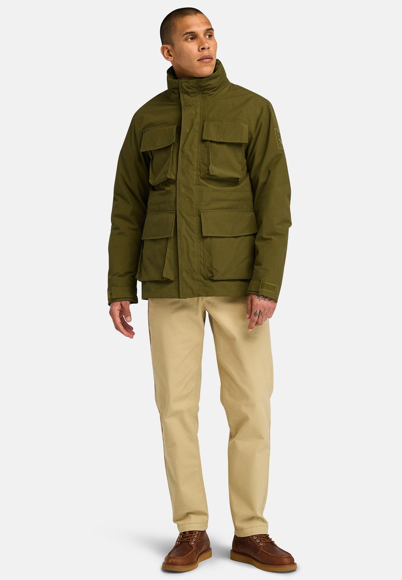 Giacca verde oliva, resistente, con colletto alto, multiple tasche frontali e taglio dritto; abbinata a pantaloni beige e scarpe marroni.