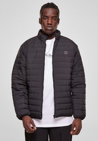 Black puffer jacket with a quilted texture, high collar, full zipper, and elastic cuffs. Worn over a white shirt, displayed against a gray background.