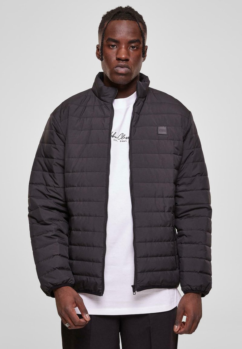 Black puffer jacket with a quilted texture, high collar, full zipper, and elastic cuffs. Worn over a white shirt, displayed against a gray background.