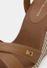 Sandale compensée marron avec des brides croisées, arborant un accent doré marqué. Brides en matériau lisse ; motif à carreaux sur le talon.
