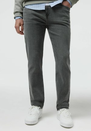 Mężczyzna ubrany w szare jeansy typu slim fit, białe trampki, jasnoniebieską koszulę w paski i zielony sweter, stoi z jedną ręką w kieszeni.