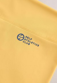 Kollane spordiriietus siledast materjalist, millel on sinine logo, mis ütleb "SELF ATHLETICS CLUB" ääre lähedal.