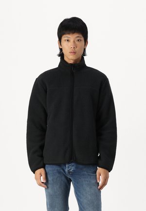 Veste en polaire noire avec un col haut, fermeture éclair et tissu texturé. Présente une coupe ajustée et des manches longues.