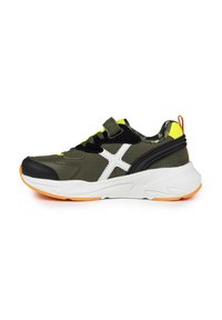 Zapatilla deportiva verde y negra con acentos blancos y amarillos, que presenta una correa de velcro, una parte superior texturizada y una suela blanca acolchada con detalles naranjas.