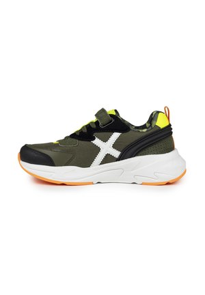 Zapatilla deportiva verde y negra con acentos blancos y amarillos, que presenta una correa de velcro, una parte superior texturizada y una suela blanca acolchada con detalles naranjas.