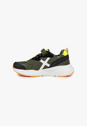 Zapatilla deportiva verde y negra con acentos blancos y amarillos, que presenta una correa de velcro, una parte superior texturizada y una suela blanca acolchada con detalles naranjas.