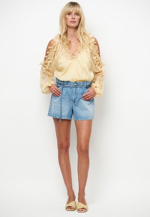 Blouse jaune clair à manches volantées, associée à un short en jean taille haute de teinte moyenne avec une ceinture à la taille. Porté avec des sandales tressées.