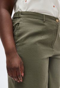 Κοντινό πλάνο ενός ατόμου που φοράει παντελόνι ελιάς με πλαϊνές τσέπες και ένα μερικώς ορατό λευκό πουκάμισο με μικρά κεντημένα σχέδια.