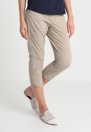 Person iført beige kortbukser og hvide vævede slip-on sko, stående med det ene ben over det andet mod en ensfarvet baggrund.