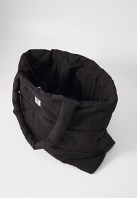 Svart, vadderad tote-väska med en strukturerad tyg, stor öppning, två handtag och ett litet logomärke på sidan. Mjukt, lätt material.