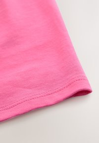 Ružová bavlnená látka s hladkou textúrou, vybavená prešitým lemom na okraji. Látka je mierne pokrčená, čo dodáva nenútený vzhľad.