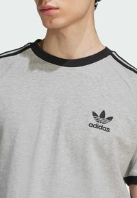 Szürke pamut póló fekete bordás kerek nyakkal, fekete háromcsíkos mintázattal a vállakon és hímzett Adidas logóval a mellkason.
