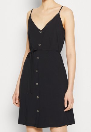 Femme portant une robe noire sans manches à boutons sur le devant, avec de fines bretelles et une ceinture à la taille, sur un fond clair uni.