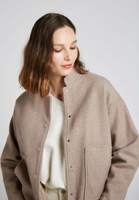Veste en mélange de laine beige avec un col montant, des fermetures à pression et une poche à l'avant. Texture douce et coupe décontractée, portée par-dessus un haut blanc.