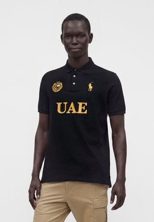 Mężczyzna ubrany w czarną koszulkę polo z żółtym napisem „UAE” i logo, w zestawieniu z jasnobrązowymi spodniami typu cargo, stojący na jednolitym białym tle.