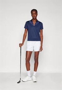 Žena oblečená v námořnickém polo, bílých šortkách a teniskách, stojí s golfovým putterem před jednobarevným pozadím.