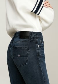 Tmavomodré džínsy z denimu s textúrovaným dizajnom, s čiernou nášivkou značky na páse a malým logom na zadnej vrecku.