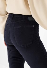 Zwarte corduroy jeans hebben een getailleerd ontwerp met achterzakken en een subtiele textuur. De tailleband toont een klein merklabel.