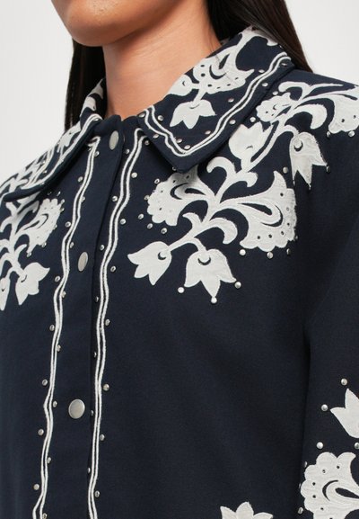 Marineblaues Jacket mit weißer Blumenstickerei, das einen großen Kragen und metallische Druckknöpfe aufweist. Texturiert mit perlenähnlichen Akzenten.
