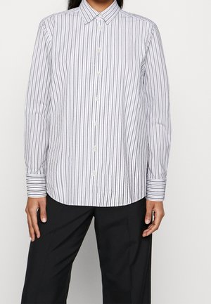 Kobieta ubrana w białą koszulę na guziki z pionowymi czarnymi paskami i czarne spodnie, stojąca z rękami swobodnie opuszczonymi wzdłuż ciała.