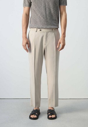 Mann trägt beige maßgeschneiderte Hose, graues kurzärmeliges Strickoberteil und schwarze Riemchensandalen, steht vor einem schlichten hellen Hintergrund.