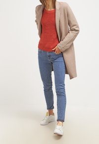 Beiger langer Mantel über einem roten strukturierten Pullover, kombiniert mit blauen engen Jeans und weißen Turnschuhen mit gelben Akzenten.