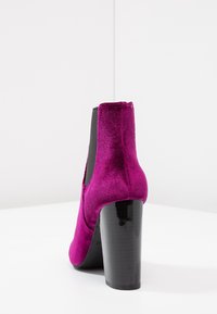 Botín de terciopelo magenta con un alto tacón de bloque negro brillante, que presenta un panel de tejido elástico en el costado para facilitar su uso.