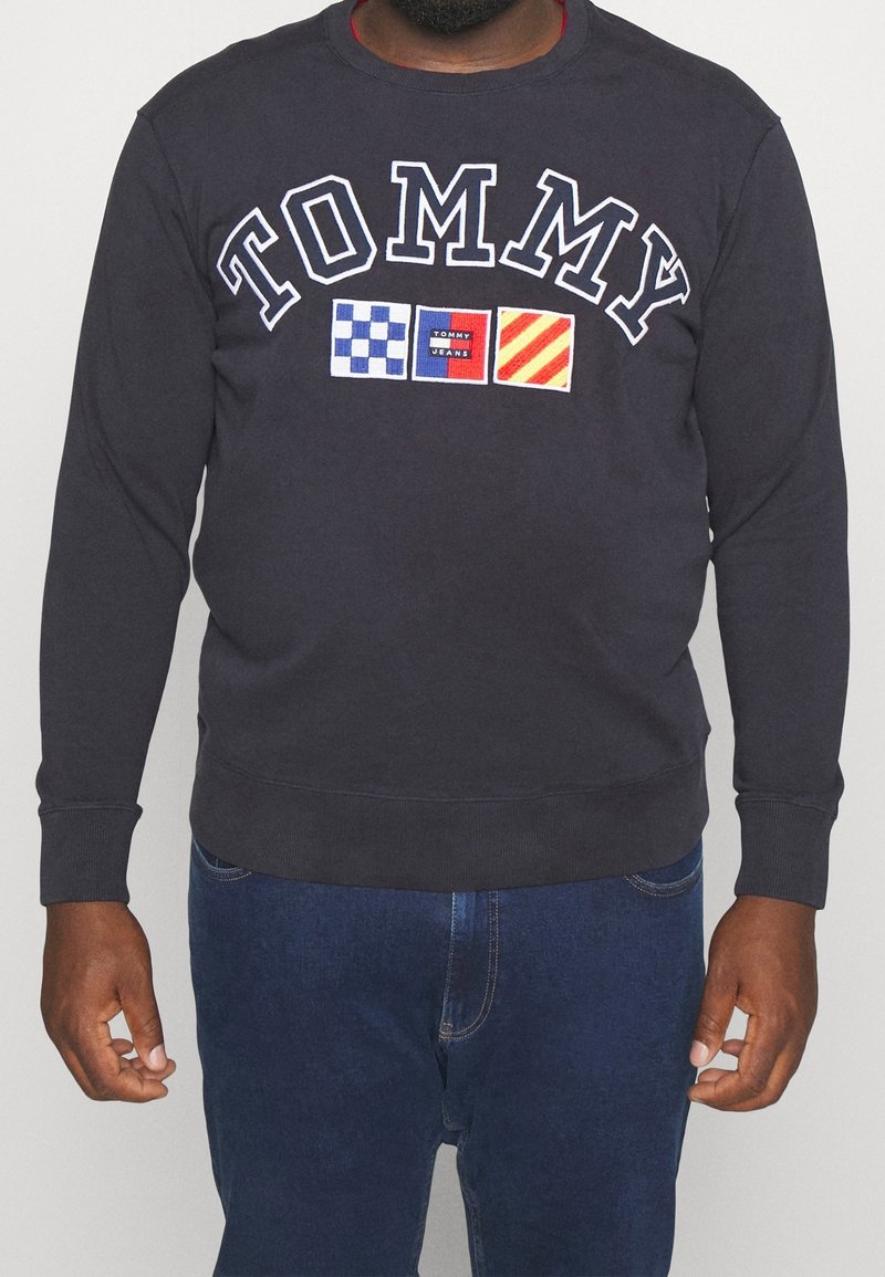 Homme portant une chemise noire à manches longues avec le texte "TOMMY" et trois logos carrés colorés sur la poitrine, associée à un jean bleu.