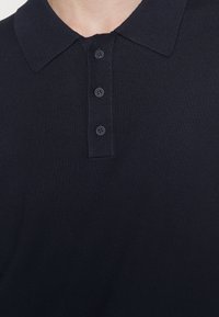 Polo shirt blu navy con maglina testurizzata, presenta un colletto classico e una patta con quattro bottoni. Dettagli di cucitura sottili lungo i bordi.
