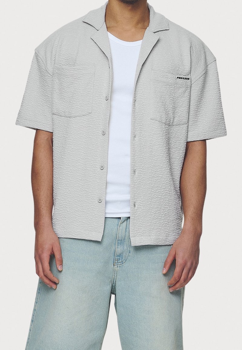 Hellgraues Kurzarm-Hemd mit Knopfleiste und strukturierter Oberfläche, ausgestattet mit zwei Taschen vorne und dem "PEGLADOR"-Logo auf der linken Seite.