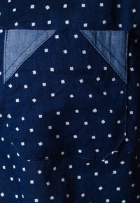 Tessuto blu navy con motivi a quadri bianchi, accentuato da una tasca in denim. La texture appare liscia con una leggera lucentezza.