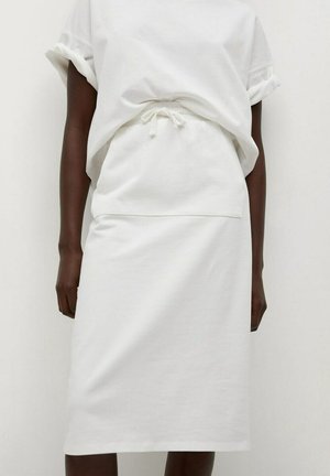 Person iført en hvid snøreskørt og en løstsiddende hvid top med korte ærmer mod en ensfarvet hvid baggrund.