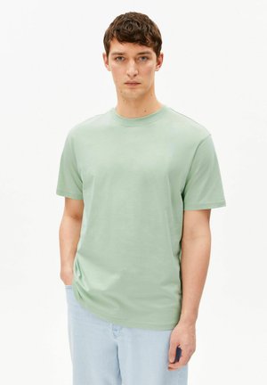 Mann trägt ein hellgrünes kurzärmeliges T-Shirt und hellblaue Jeans, steht mit einer Hand in der Tasche vor weißem Hintergrund.