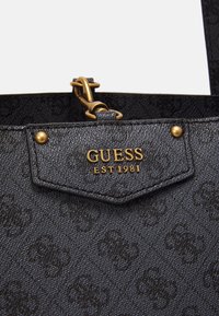 Sort tekstureret håndtaske med guldmetall detaljer. Har et logo patch prægning med "GUESS EST 1981" og dekorative nitter.