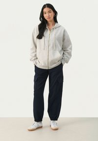 Jasnoszara bluza rozpinana z kapturem i sznurkami, z przednimi kieszeniami, noszona z granatowymi spodniami cargo. Model stoi z rękami w kieszeniach, w białych sneakersach.