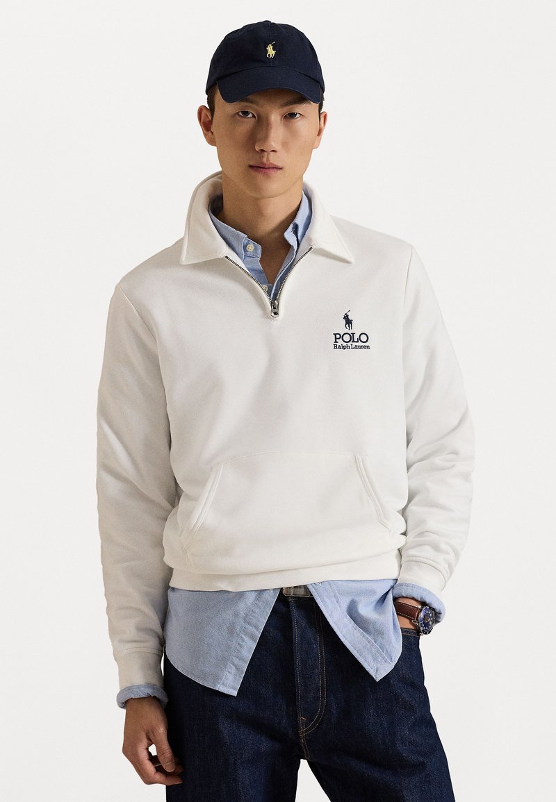 Valge fliispullover poolsärgiga, eesmine tasku ja tumesinine brodeeritud logo. Kantud helesinise kraega särk ja tumedad teksad.